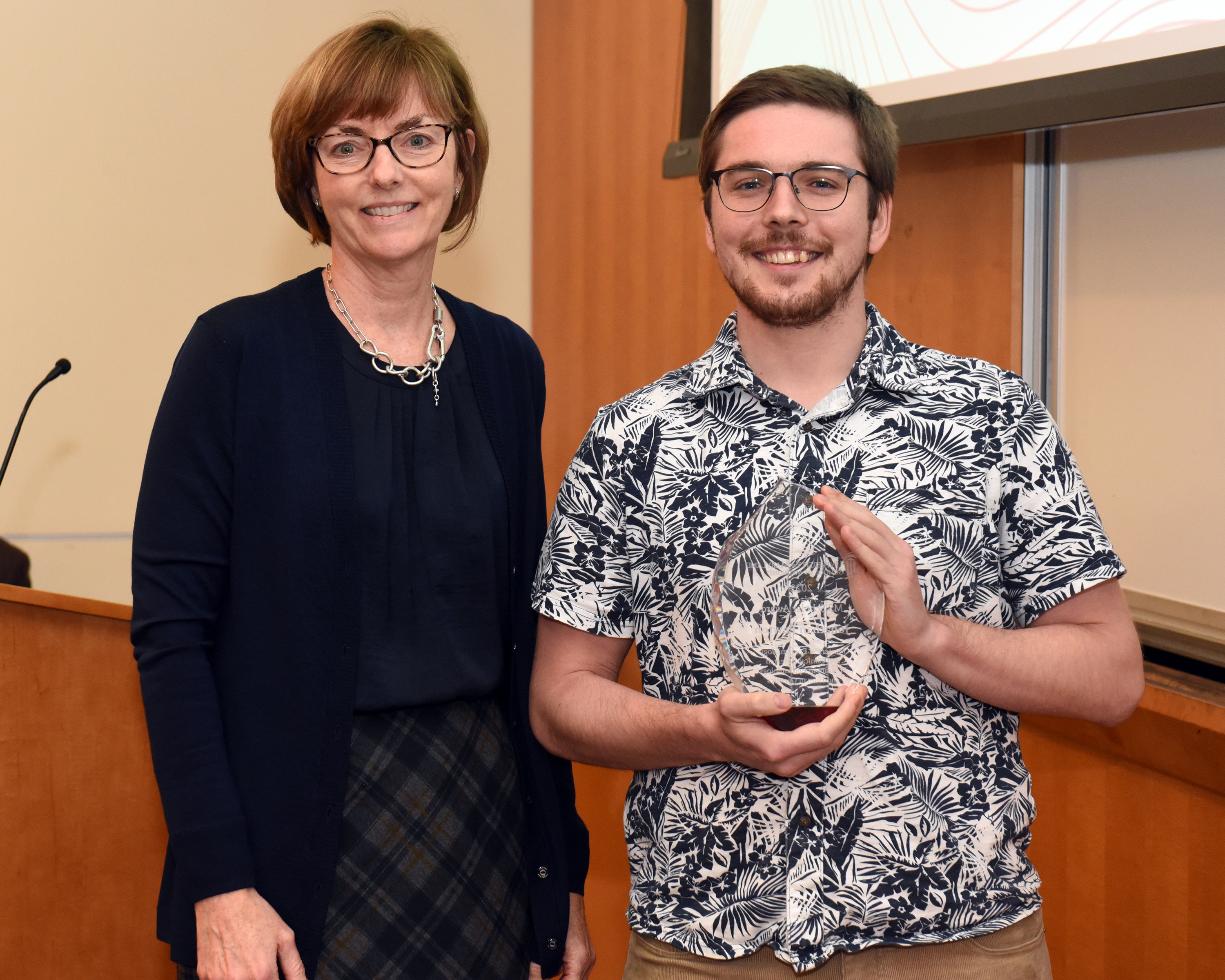 Dr. Eileen Barry is a white woman with short brown hair smiling wearing a black cardigan, blouse, and skirt standing next to Jonathan Lawton, a white man with brown hair, glasses, and a beard, wearing a black and white floral shirt. He is holding his award.