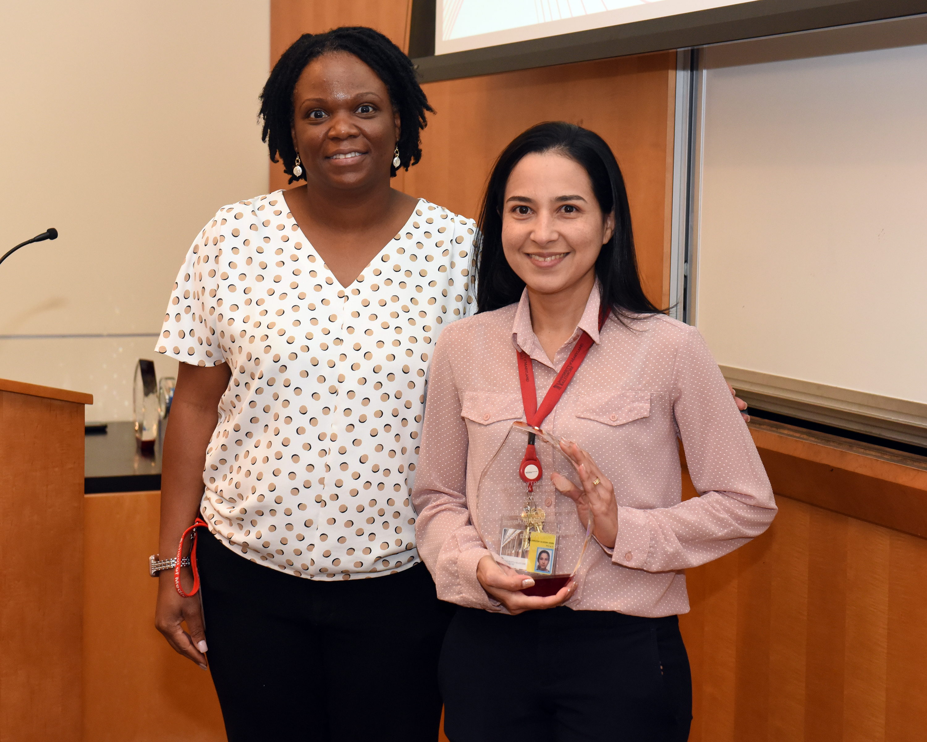 Dr. Tanya Webb is a Black woman with black curly hair, wearing a black speckled white blouse. She is standing next to Daiana  Cardoso Oliveira Viera who is a Latina woman wearing a red and white blouse, smiling and holding her award.
