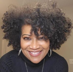 Photo of Sharron Graves. DBA, a Black woman with curly hair. She is smiling and wearing white dangling earrings and a white blouse.