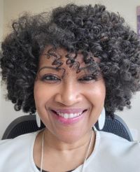 Photo of Sharron Graves. DBA, a Black woman with curly hair. She is smiling and wearing white dangling earrings and a white blouse.