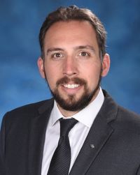 Photo of Dr. Phil Iffland. He is a white man with a brown goatee and short hair. He is wearing a white dress shirt, black tie, and black suit jacket. He is smiling against a blue background
