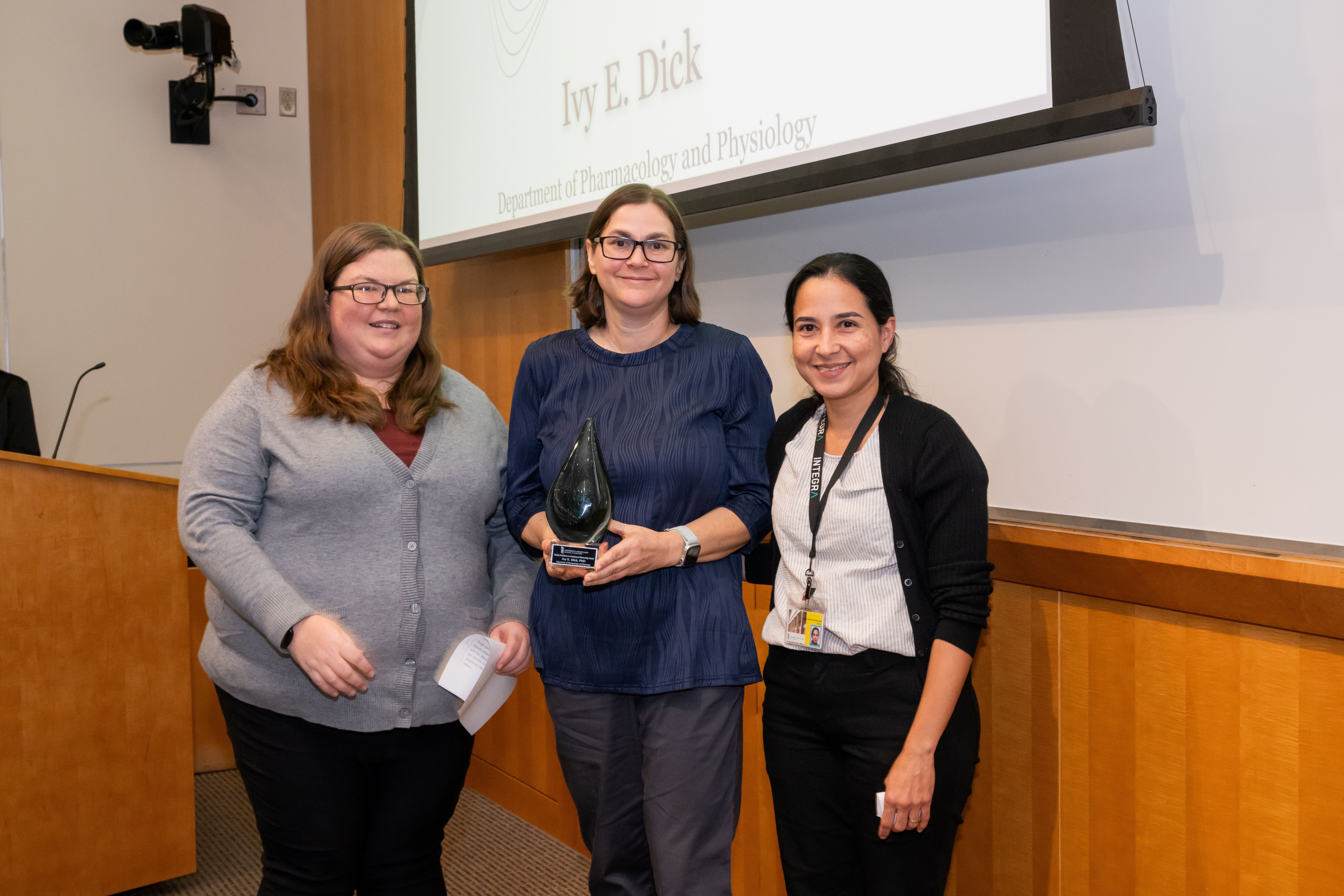 Dr. Audra Kramer, Dr. Ivy Dick, and Dr. Daiana Vieira are standing next to each other in front of a presentation screen. Dr. Dick is holding a cone shaped crystal award.