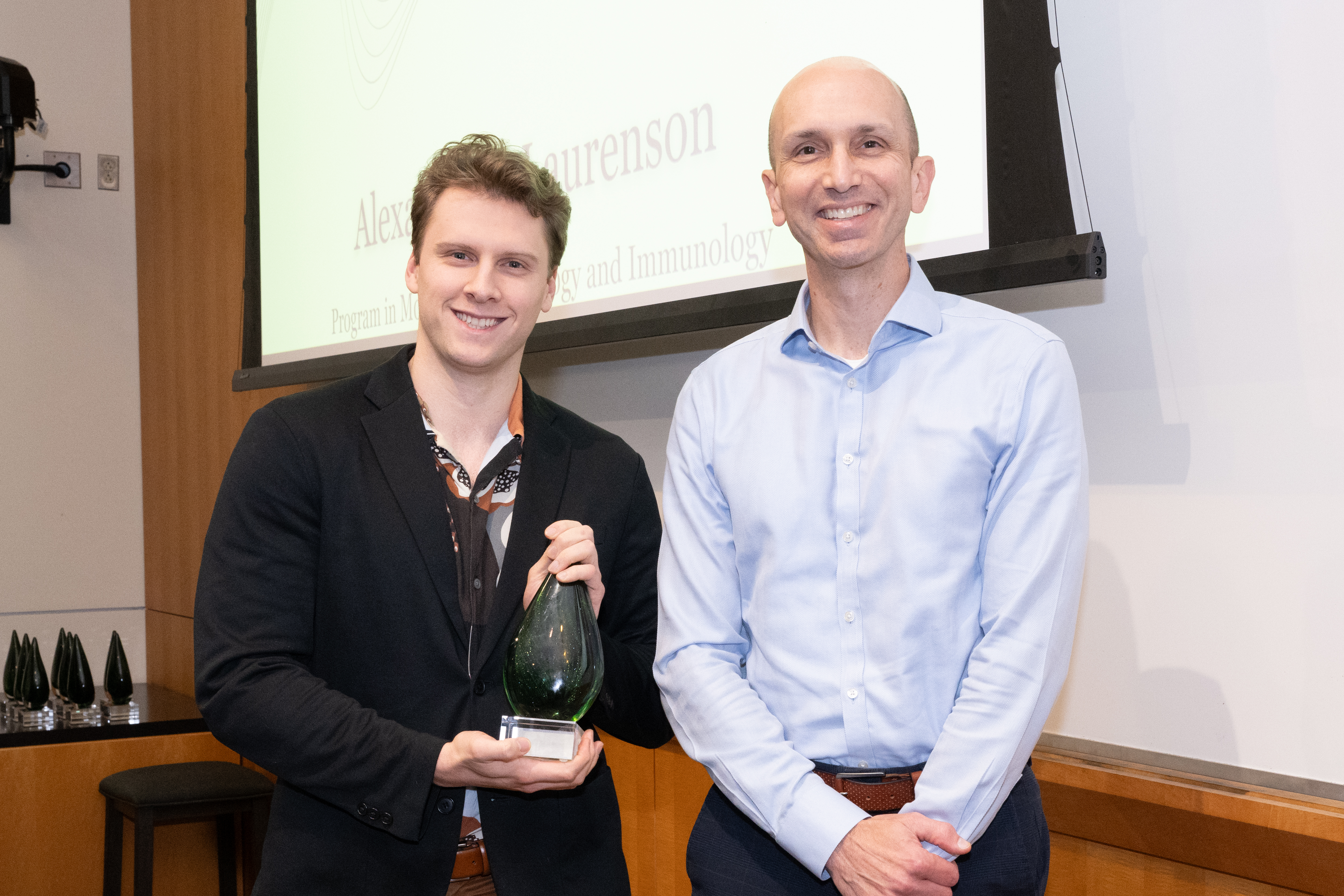 Alexander Laurenson and Dr. Matthew Laurens are standing next to each other in front of a presentation screen. Alexander is holding a cone shaped crystal award.