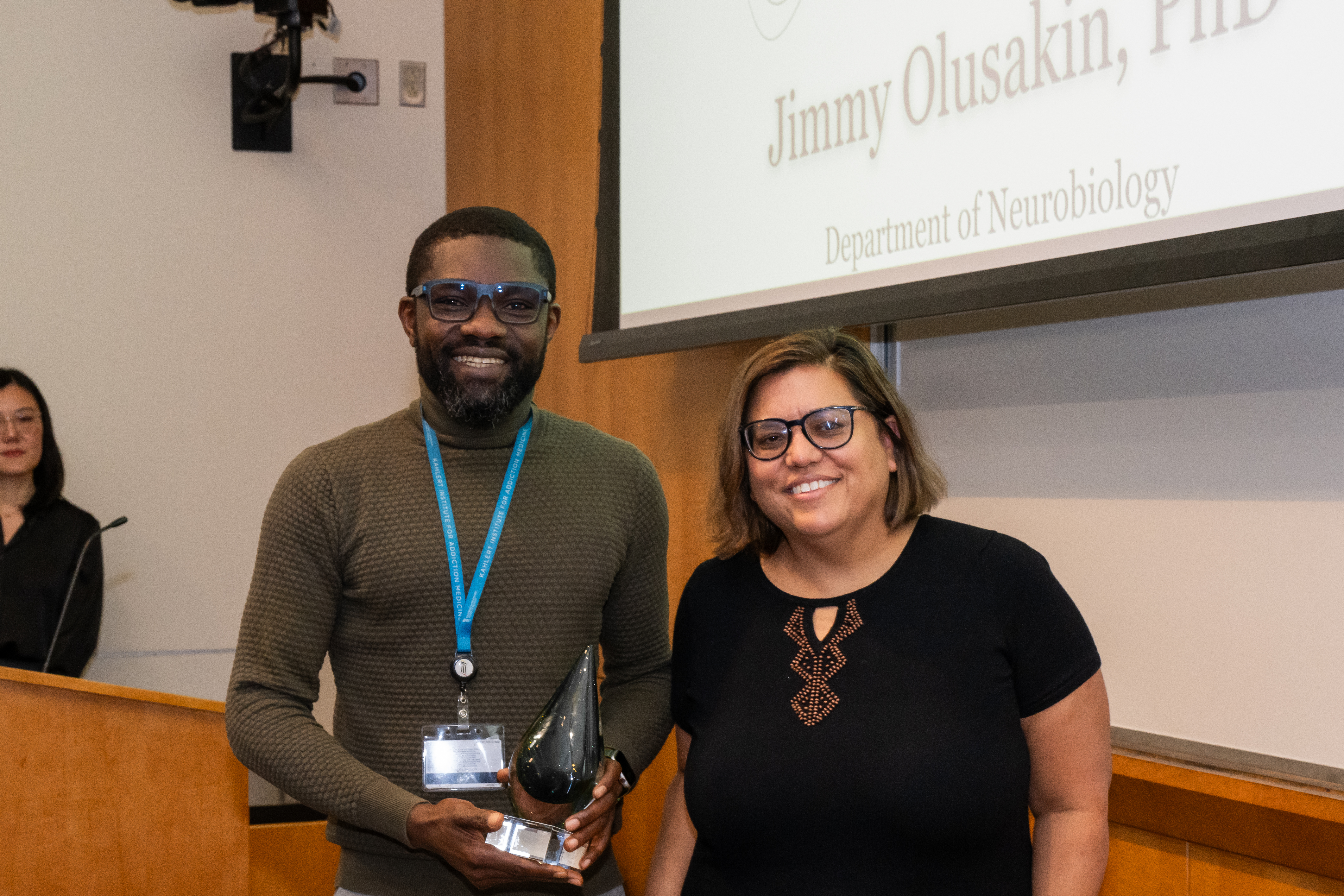 Dr. Jimmy Olusakin and Dr. Mary Kay Lobo are standing next to each other in front of a presentation screen. Dr. Olusakin is holding a cone shaped crystal award.