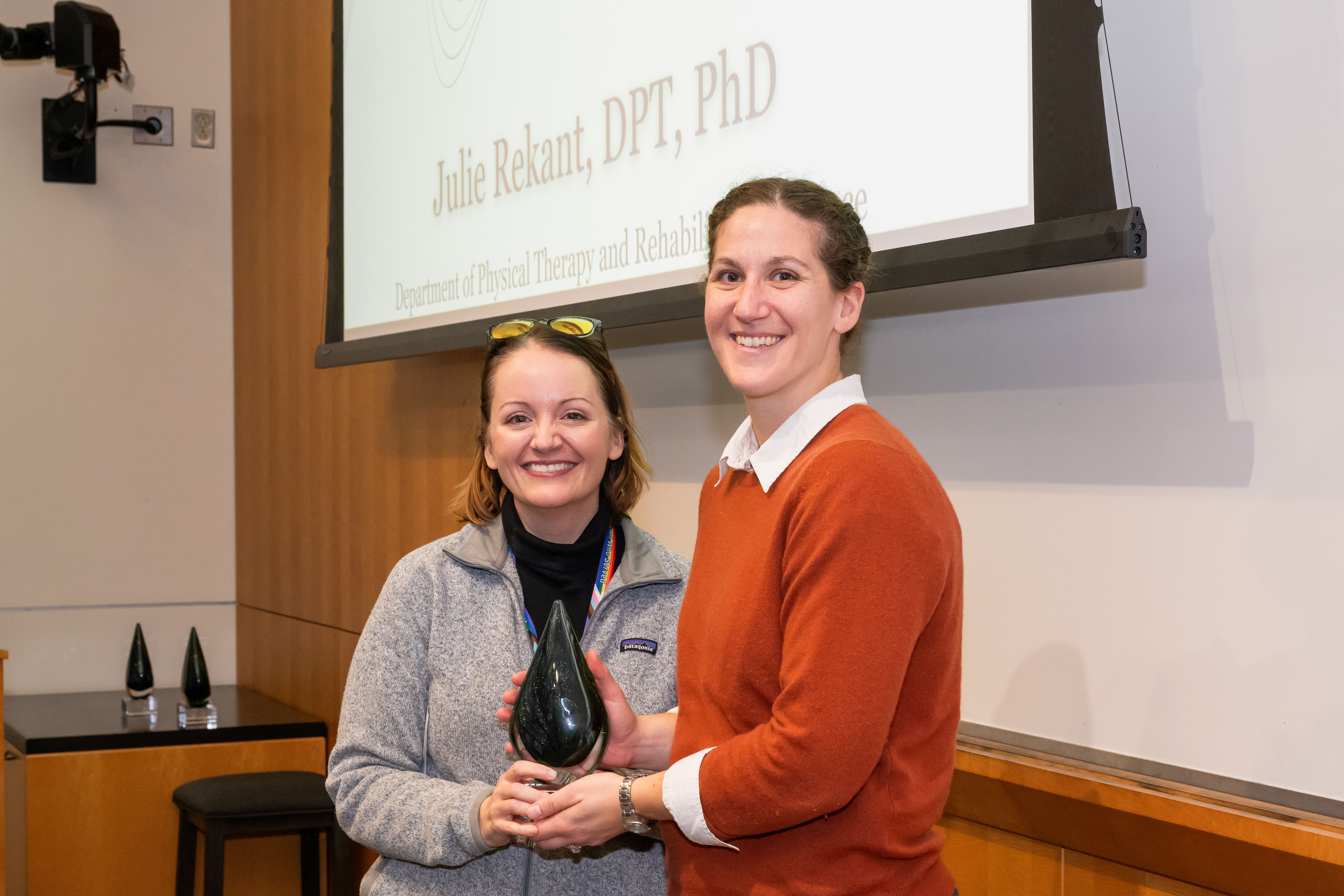 Dr. Julie Rekant and Dr. Odessa Addison are standing next to each other in front of a presentation screen. Dr. Rekant is holding a cone shaped crystal award.