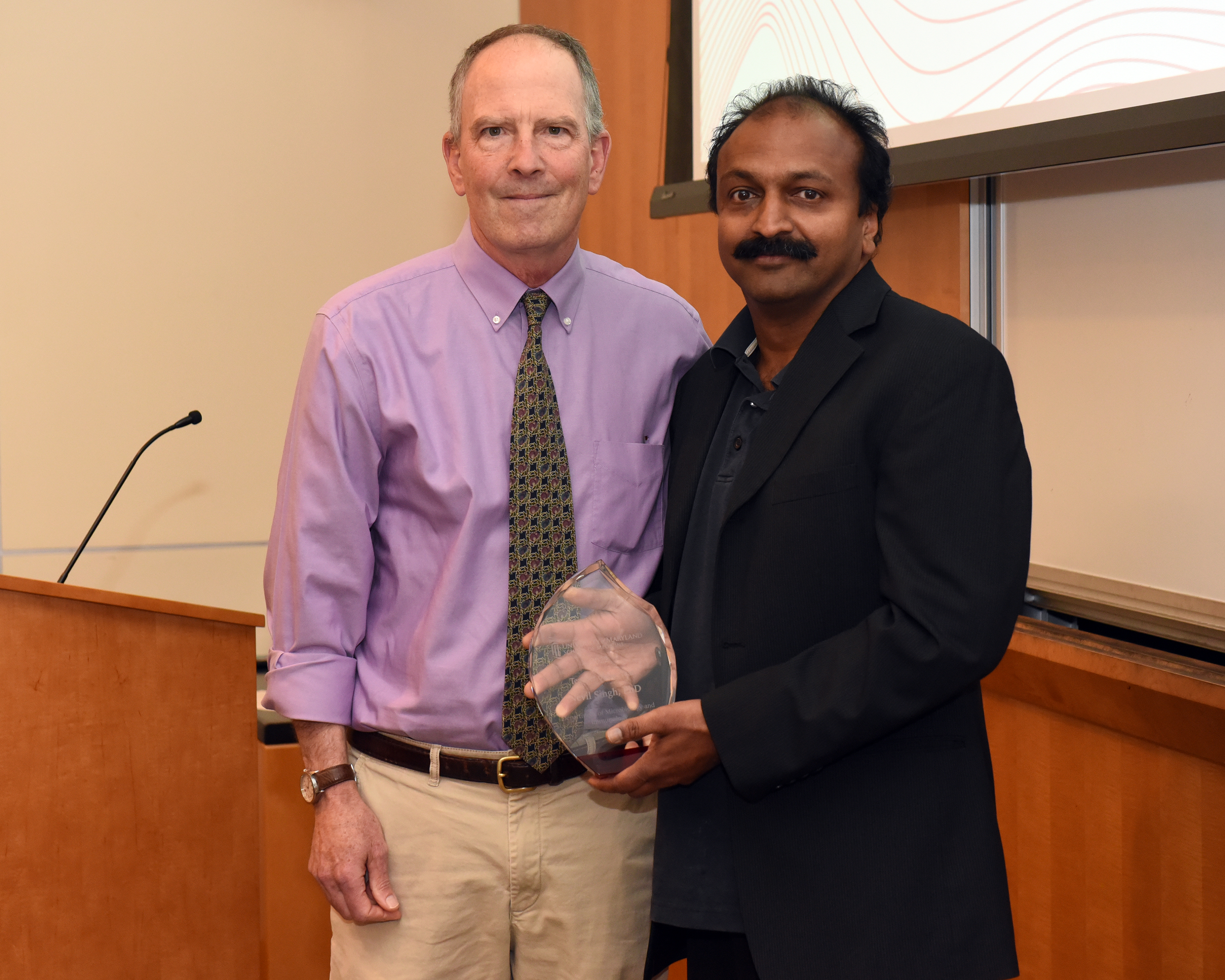 Dr. Bret Hassel, a bald white man wearing a purple shirt and black tie, is standing next to Dr. Nevil Singh, an Indian man with black hair and a mustache wearing a black suit and black shirt holding his award and smiling.