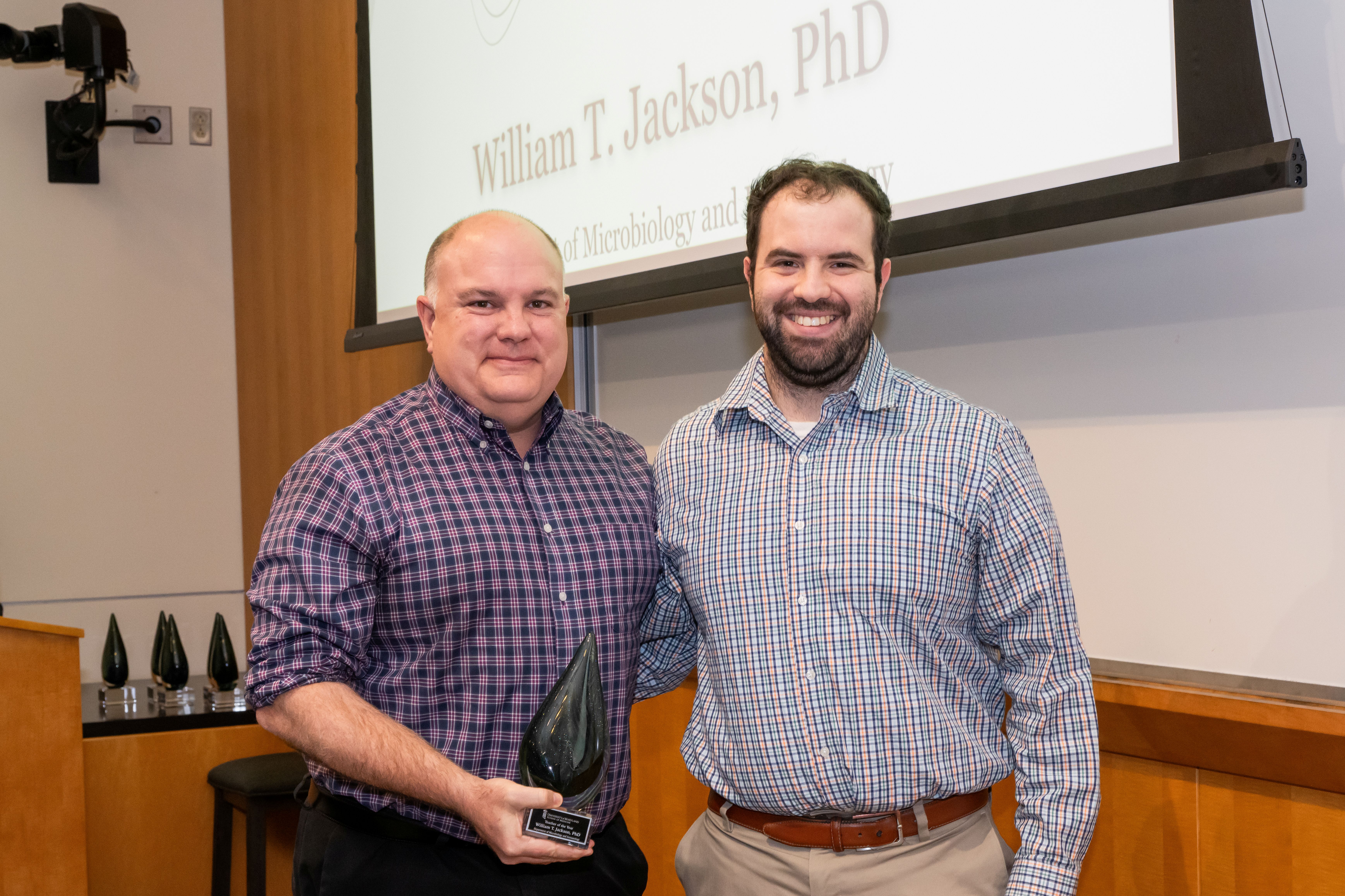 Dr. William Jackson and Noah Pollack are standing next to each other in front of a presentation screen. Dr. Jackson is holding a cone shaped crystal award.