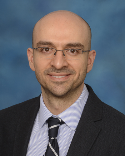 Photo of Alex Poulopoulos, PhD in a black suit, light blue shirt, and black tie, in front of a blue background