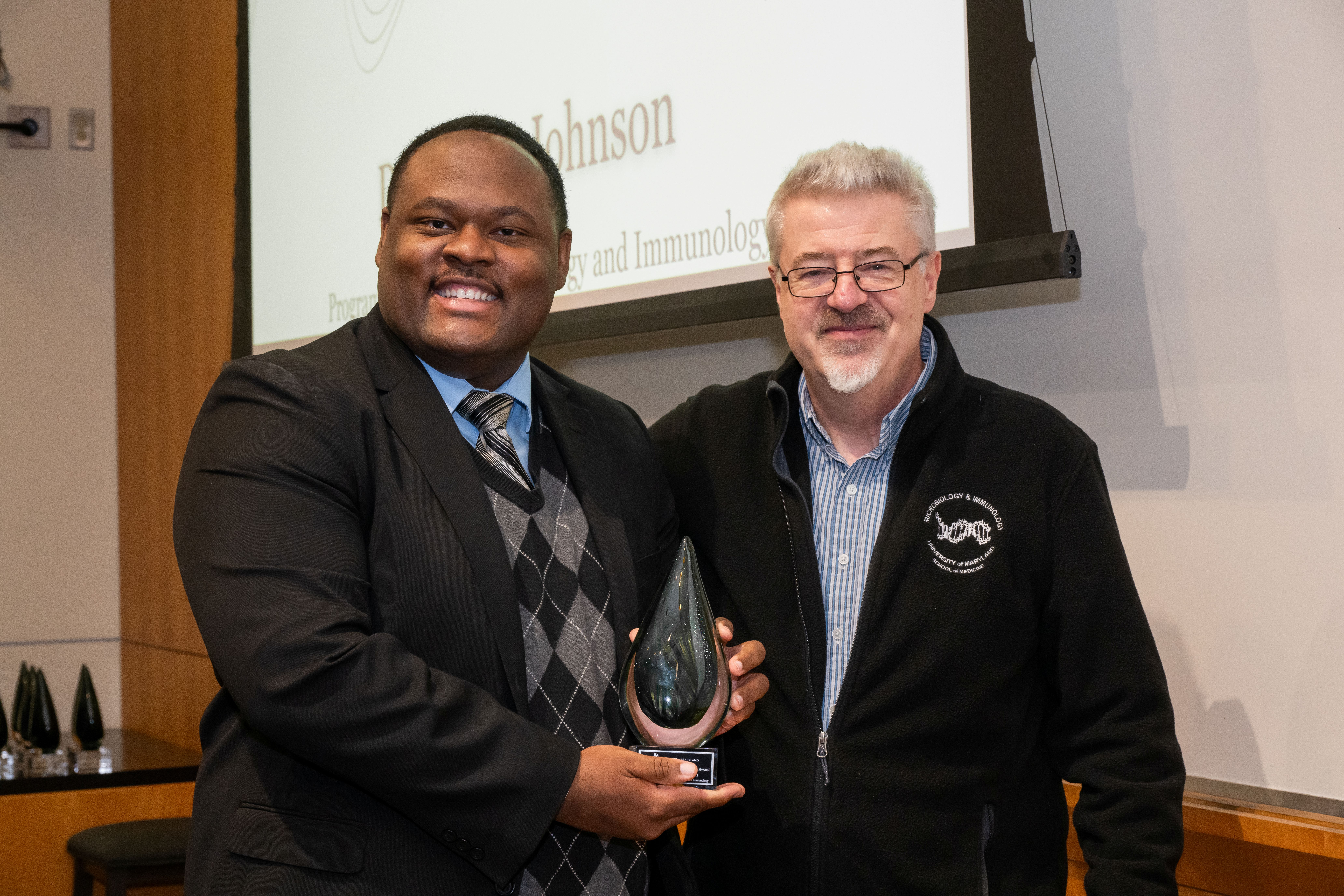 Da'Kuawn Johnson and Dr. Nicholas Carbonetti are standing next to each other in front of a presentation screen. Da'Kuawn is holding a cone shaped crystal award.