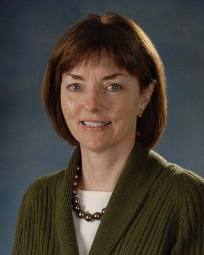 Photo of Eileen Barry. She is wearing a green blazer, white shirt, green beaded necklace, and smiling in front of a blue background.